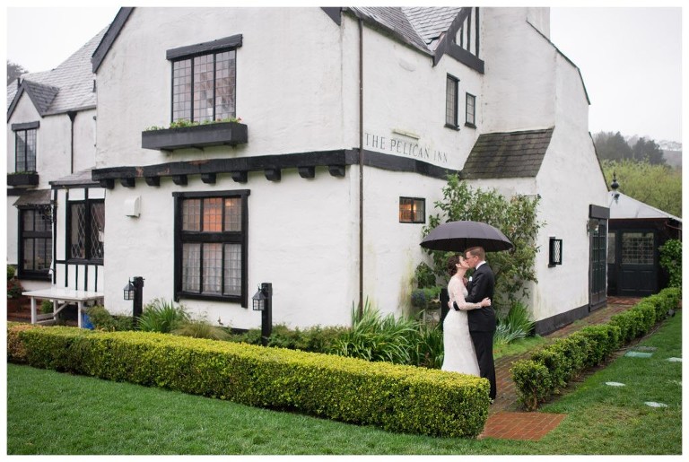 A bride and a groom kiss under umbrellas in the rain at the Pelican Inn.